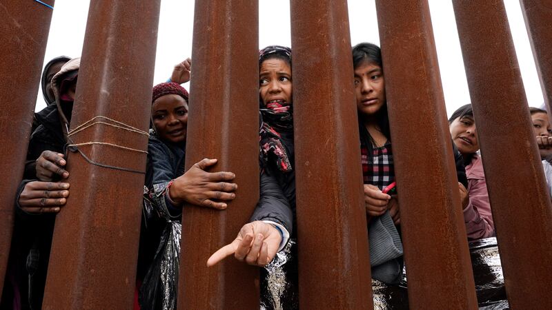 Migrants reach through a border wall for clothing handed out by volunteers, as they wait...